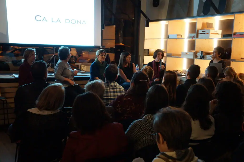 The speakers seated beneath a screen showing the title Ca la Dona during the presentation