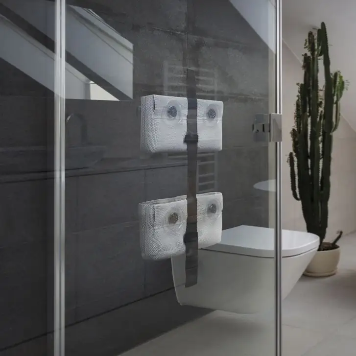 TAPE SHOWER strap with two grey pouches fixed with suction cups to the glass of a shower screen, in a bathroom with dark walls