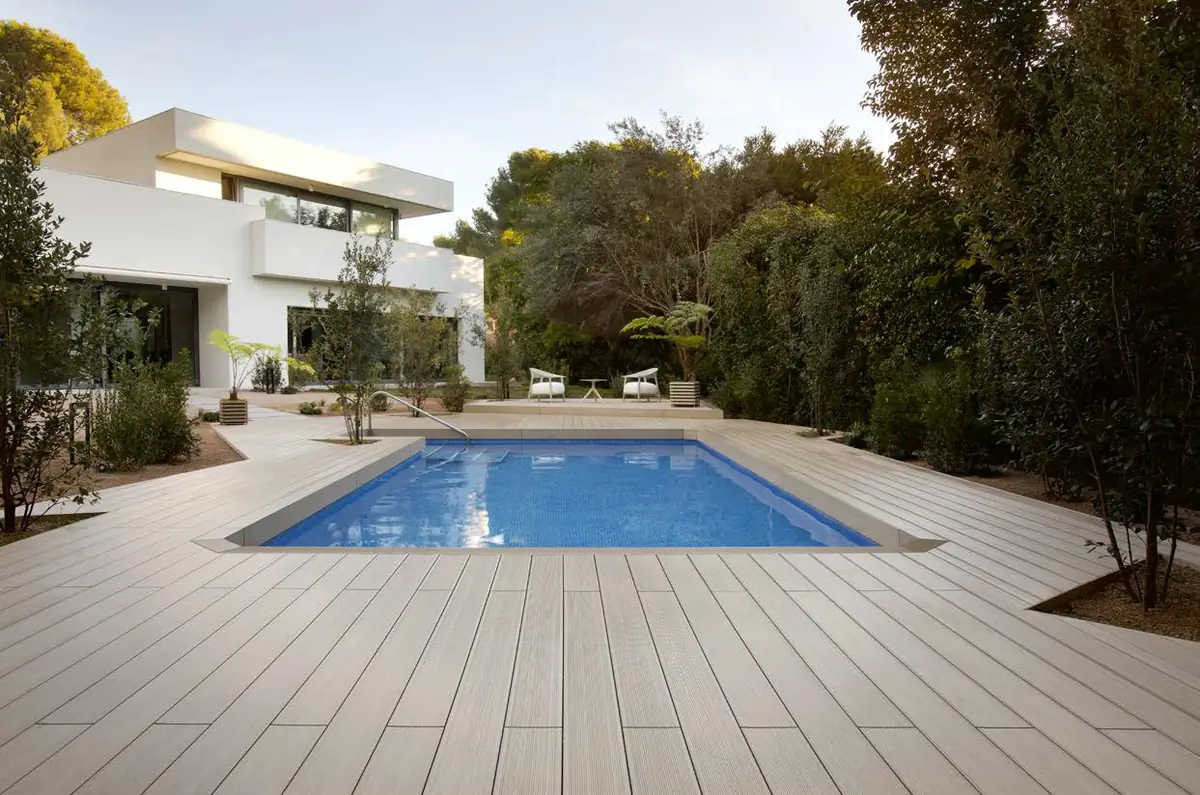 Piscina rodeada de una amplia tarima exterior de tono claro junto a una vivienda blanca