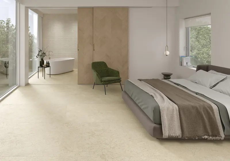 Bedroom with a light stone-effect porcelain floor and, in the background, a bathroom with a freestanding bath