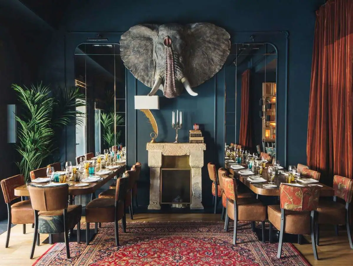 Dining room with dark blue walls, an elephant head above the fireplace, upholstered chairs and rust-coloured velvet curtains