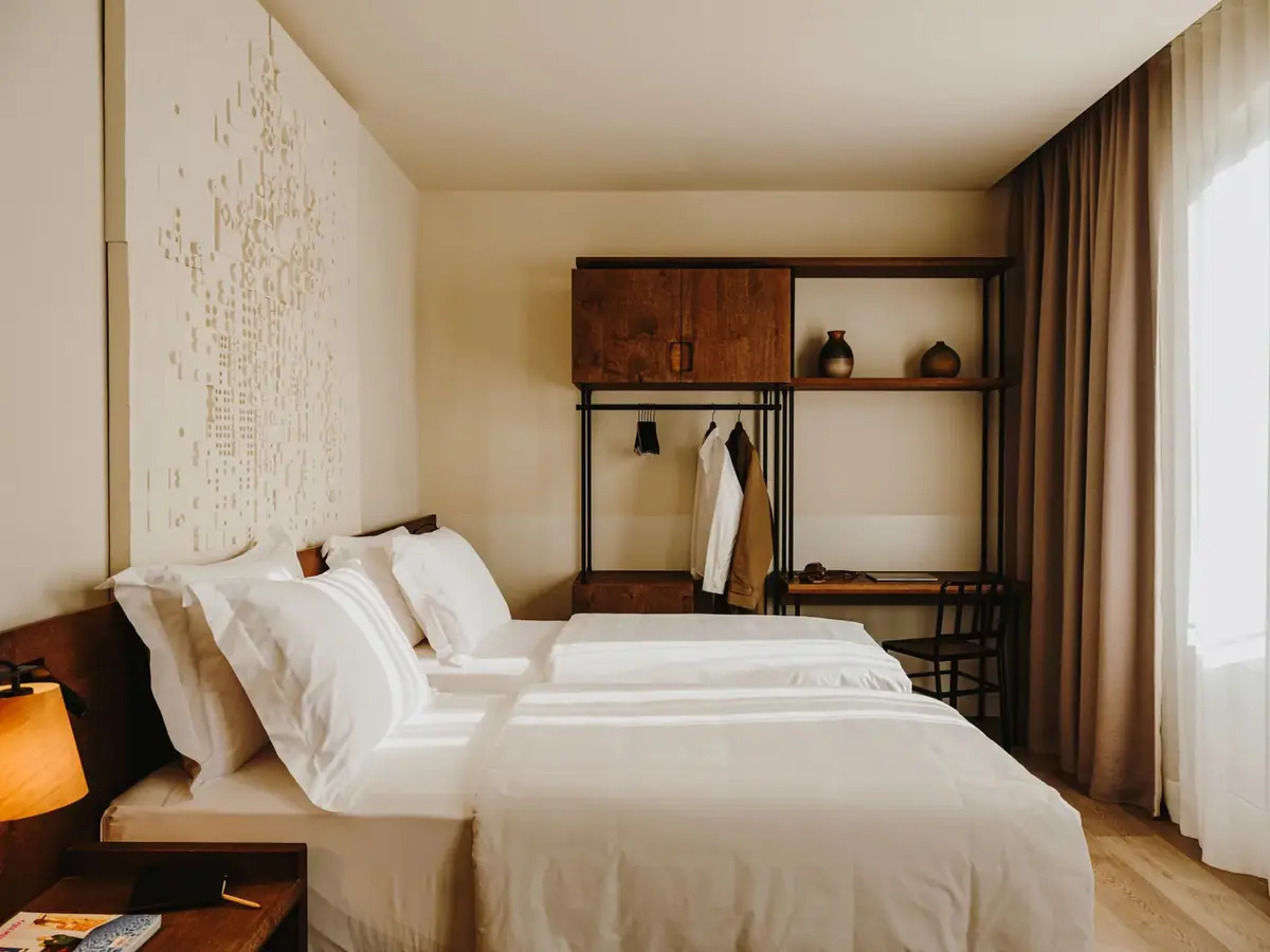 Room with two beds made up in white linen, taupe curtains, sheer curtains and an open timber unit