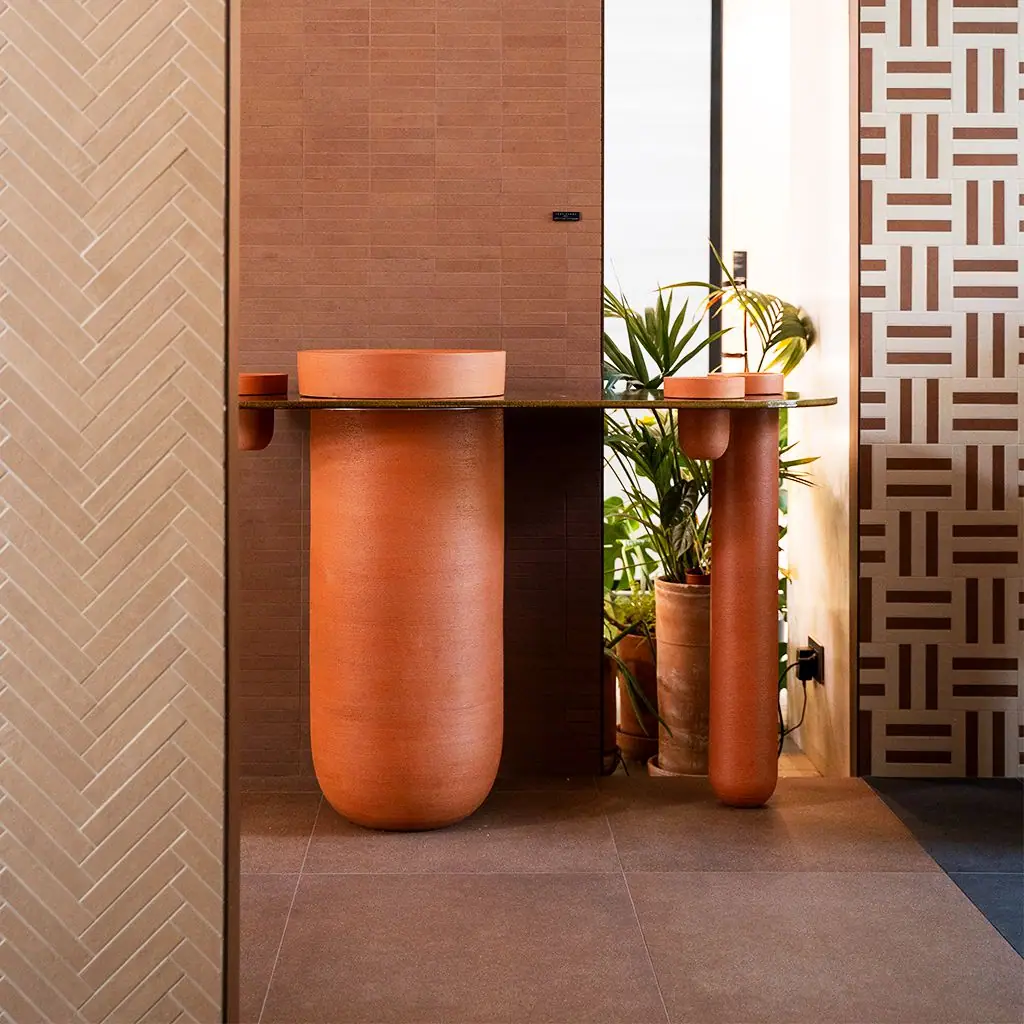Terracotta-coloured ceramic washbasin on a large column, with a dark top and a second, slimmer column, against a brick wall