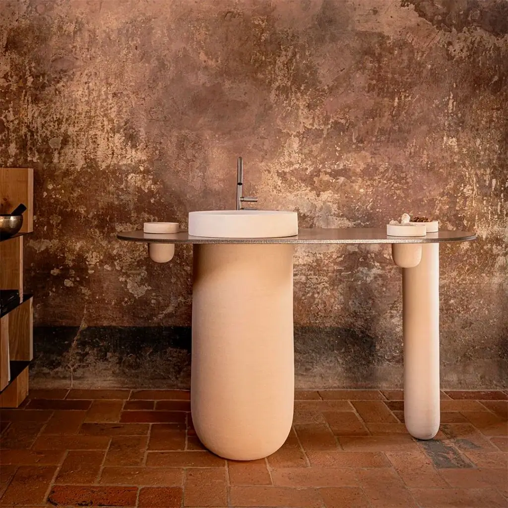 White washbasin on a sand-coloured ceramic column, with a metal top and side containers in front of an old wall