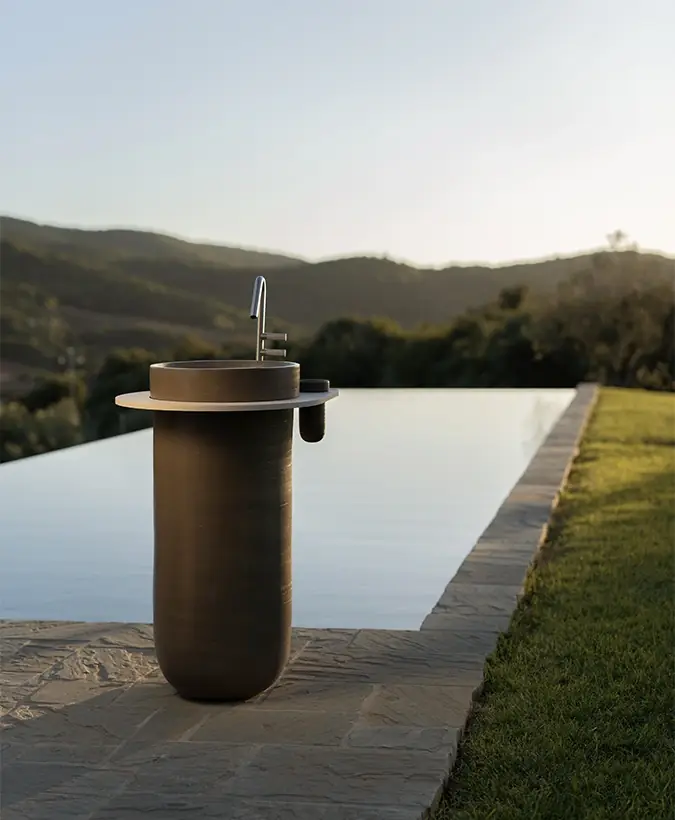 Dark ceramic column washbasin with a round top beside an infinity pool, with hills in the background