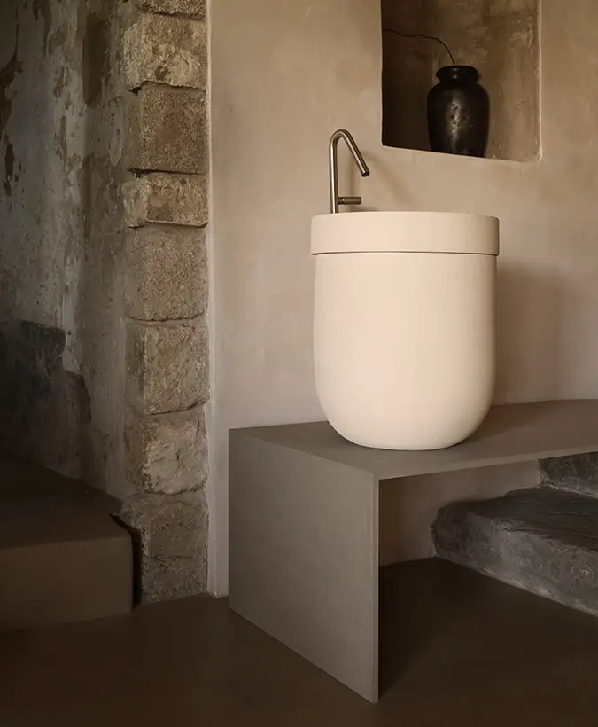 Tall light ceramic washbasin on a low bench with a steel tap, beside a stone wall