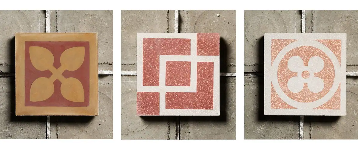 Three decorated cement tiles resting on a grey surface: a cementina with yellow leaves and two red and white graniglie