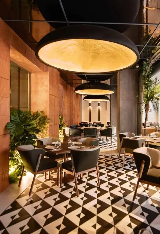 Comedor de restaurante con grandes lámparas colgantes, sillas tapizadas en gris y suelo de baldosas geométricas en blanco y negro.