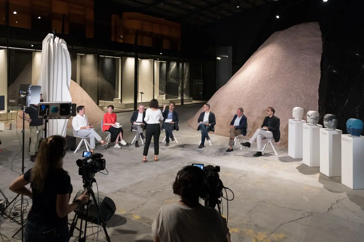 Speakers seated in a semicircle in the COMPAC showroom while two operators film them with video cameras.