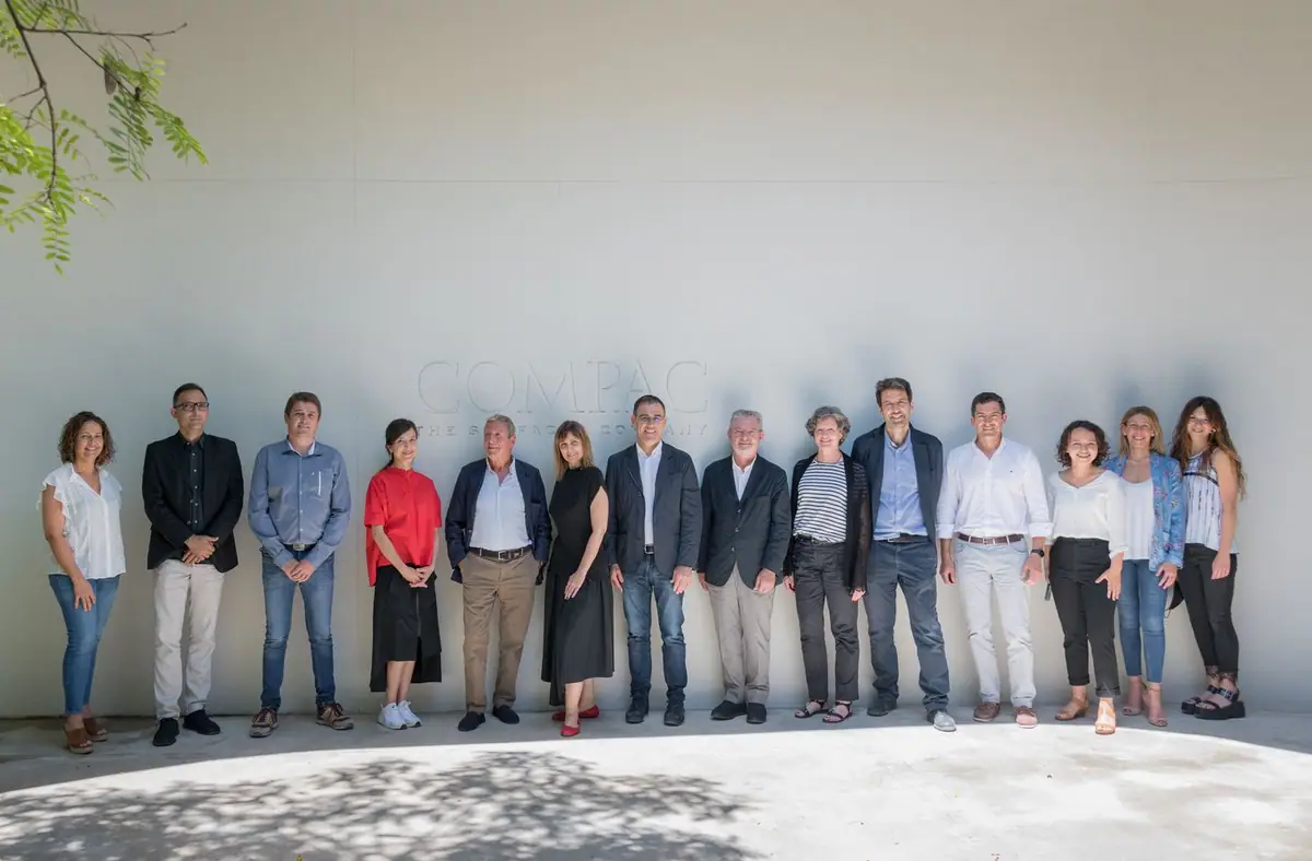 Group of people posing in a row in front of a pale wall with the COMPAC sign.