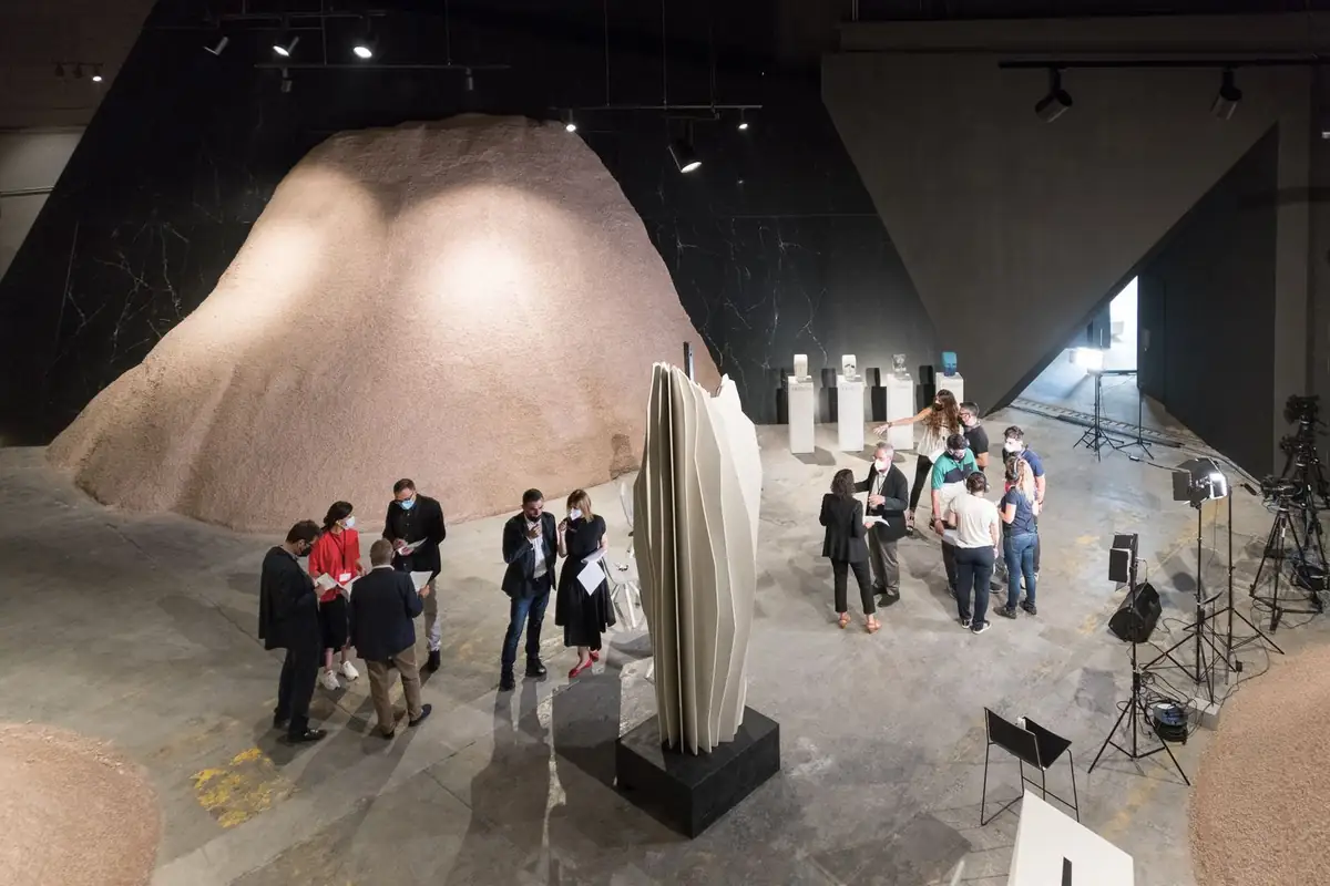 Overhead view of the COMPAC showroom with groups of people talking next to a folded white sculpture, mounds of granular material and filming lights.