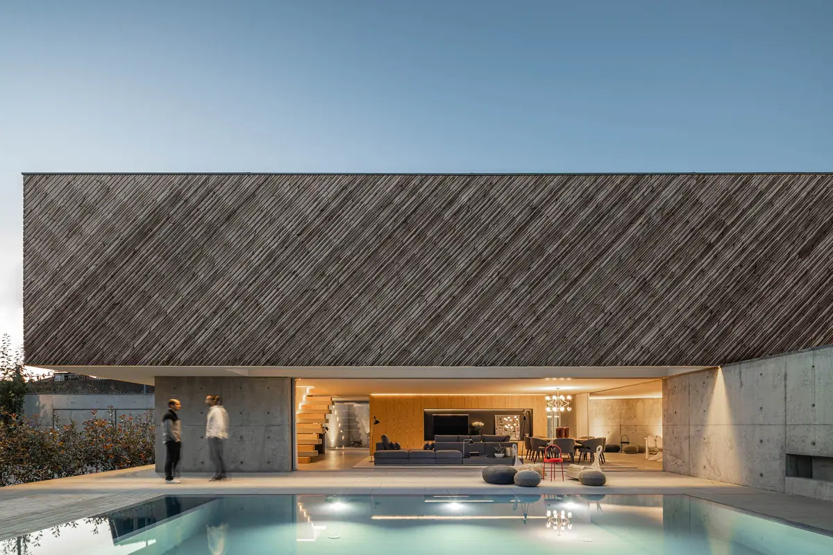 House with a large upper volume clad in diagonal timber battens above an open ground floor and a swimming pool