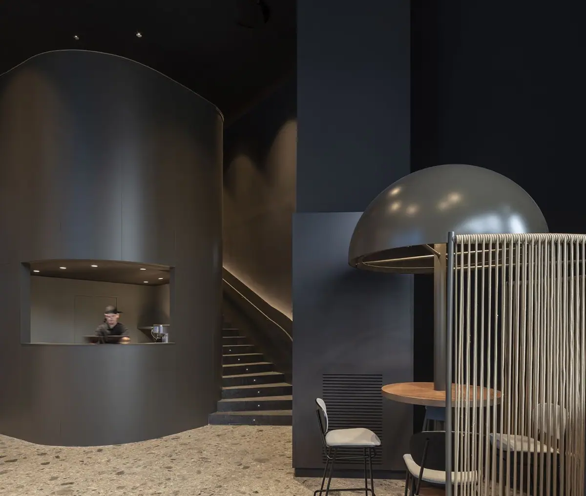 Dark curved volume with a service hatch, a staircase beyond and a high table beneath a metal dome surrounded by ropes