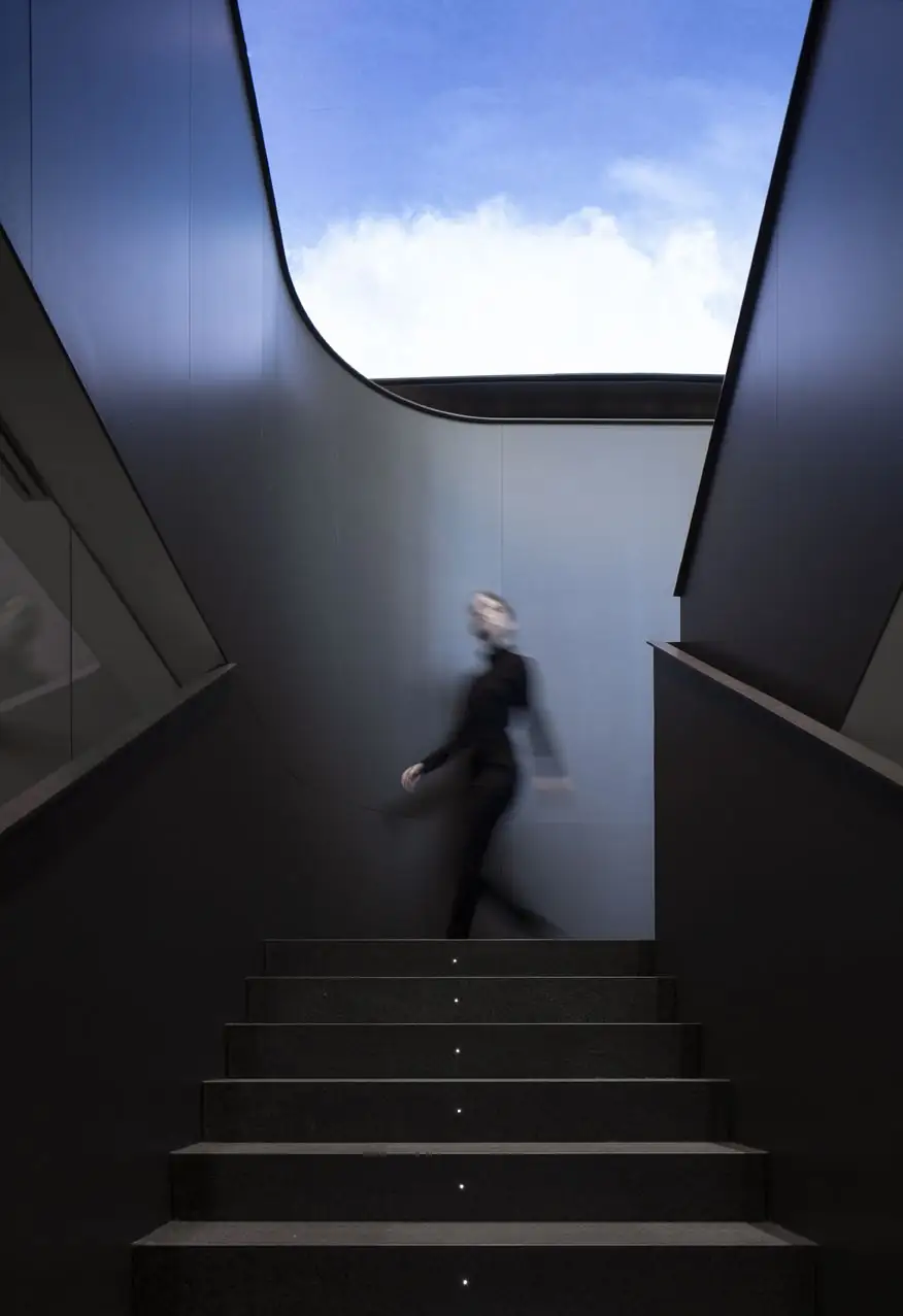 A person climbing a dark staircase between curved walls, with a sky screen above