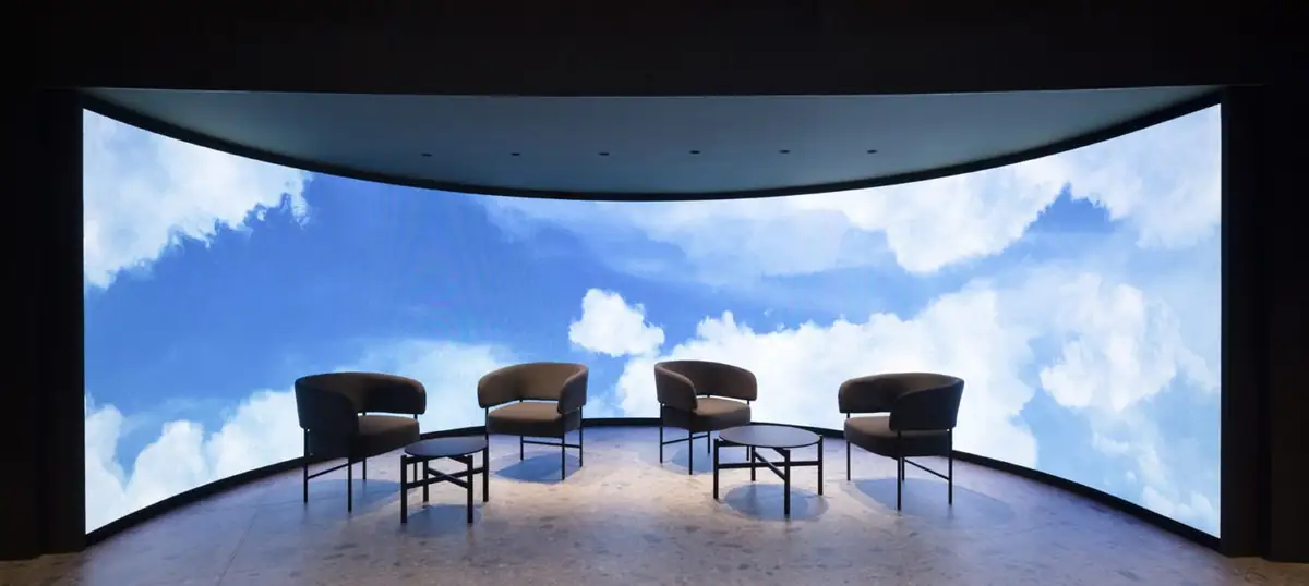 Four armchairs and two low tables in front of a large curved LED screen showing sky and clouds