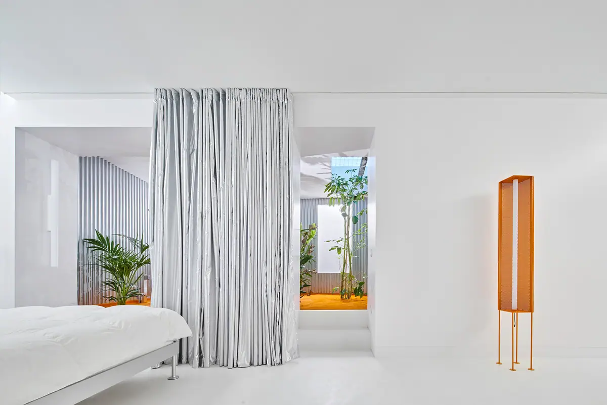 White bedroom with a silver curtain and, in the background, the interior courtyard with tropical plants and orange turf