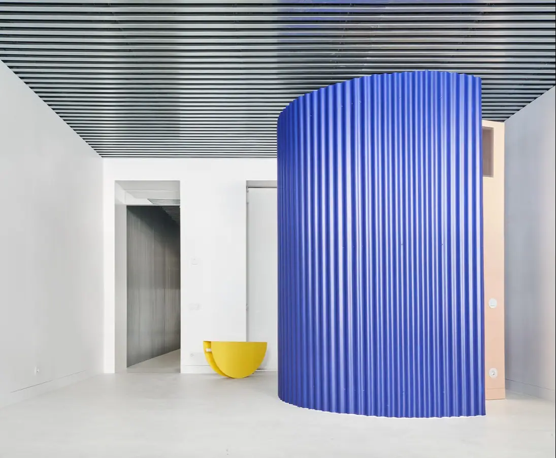 Cylindrical volume in blue corrugated sheet metal beneath a ceiling of metal slats, with a yellow piece of furniture against the wall