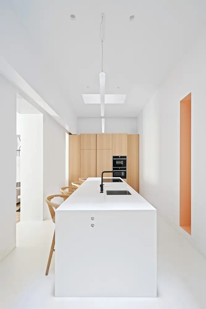 White kitchen with a long island, wooden stools, light wood cupboards and an orange niche in the wall