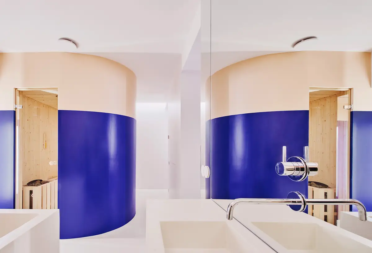 Bathroom with a white washbasin facing a curved blue and beige volume housing a wooden sauna