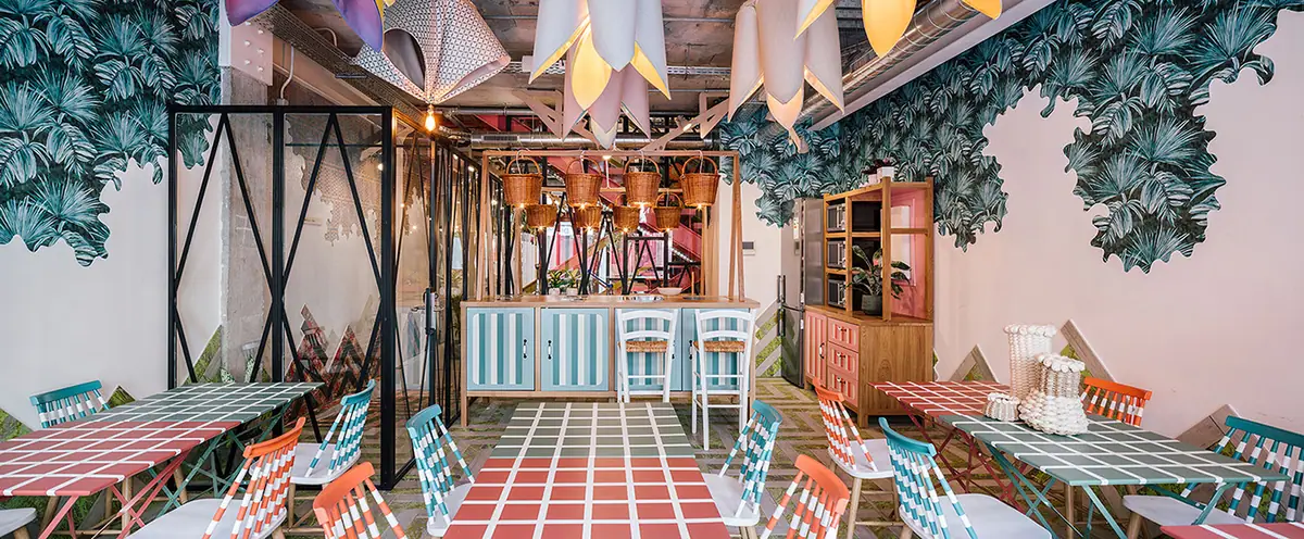 Canteen with colourful checked tables, striped chairs, leaf murals, flower-shaped lamps and baskets hanging above the bar