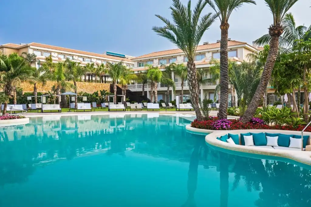 Lake-style pool among palm trees with a chill-out peninsula of white and turquoise sofas in front of the hotel buildings