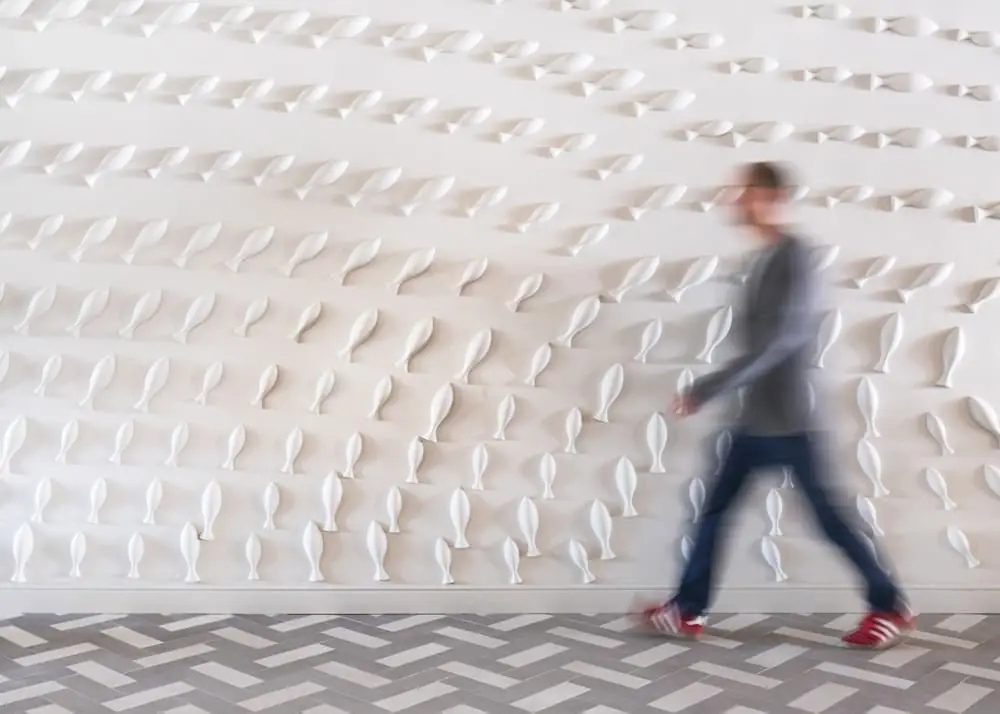 White wall with dozens of plaster fish in relief and a person walking in front