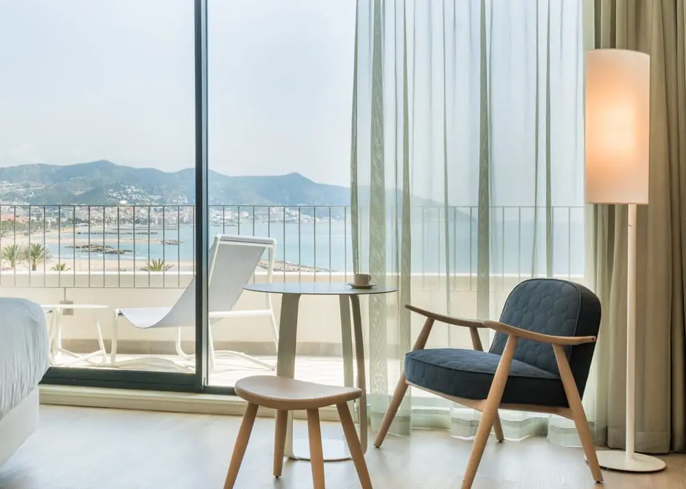 Room with an upholstered armchair, a round table and a large window opening onto a terrace with sea views