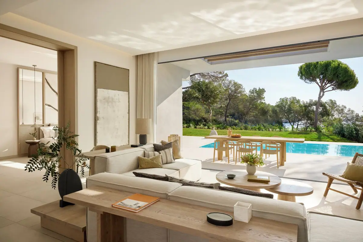 Salón de un bungalow con sofá claro, mesa de madera y ventanal abierto a la piscina y al jardín