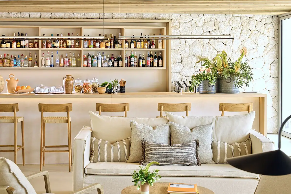 Barra de bar con estanterías de madera, muro de piedra y sofá con cojines de tonos claros