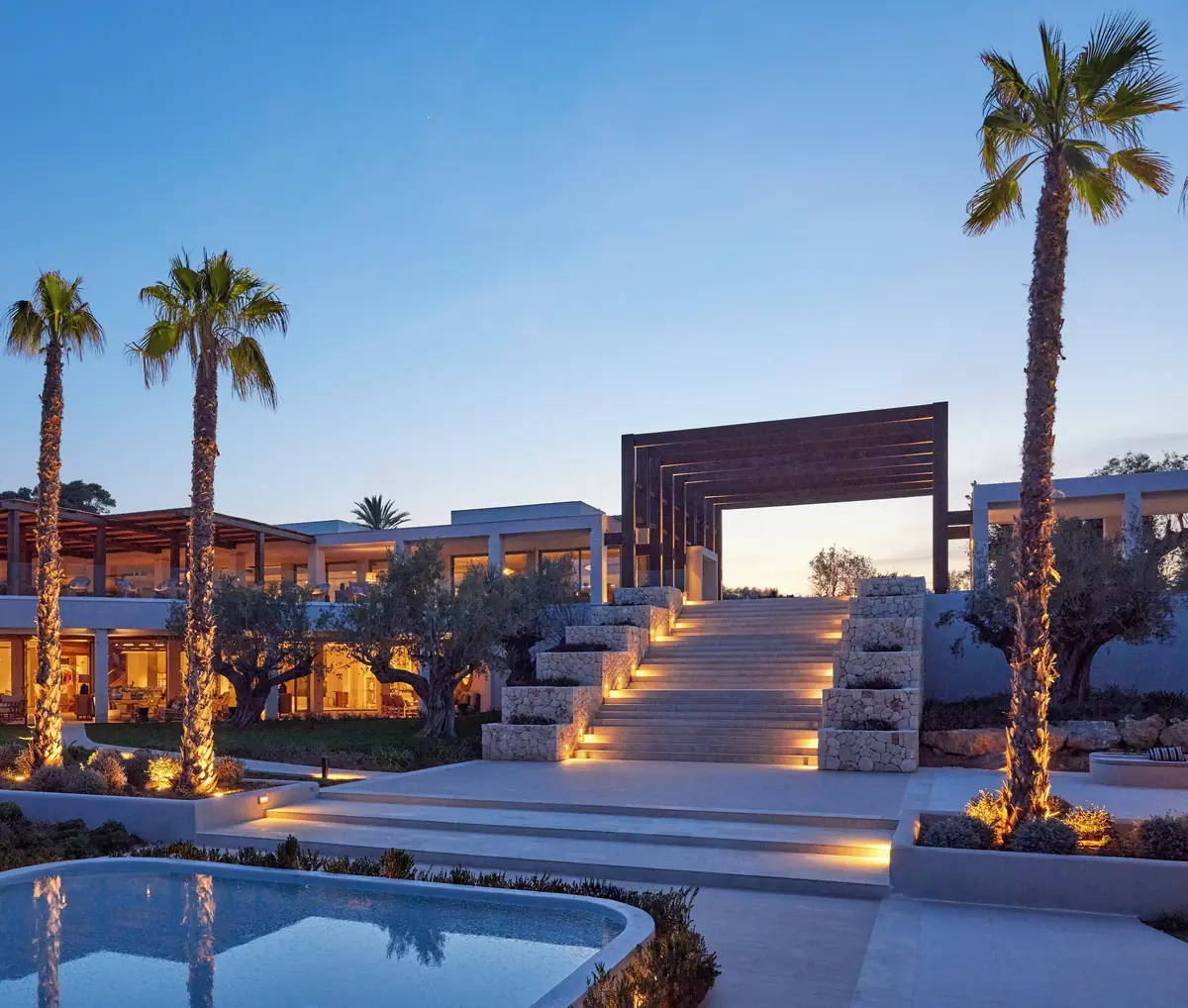 Acceso principal con escalinata iluminada, pórtico de madera, palmeras y piscina a la hora azul