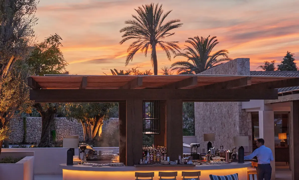 Barra exterior del lobby bar bajo una pérgola de madera al atardecer, con palmeras al fondo