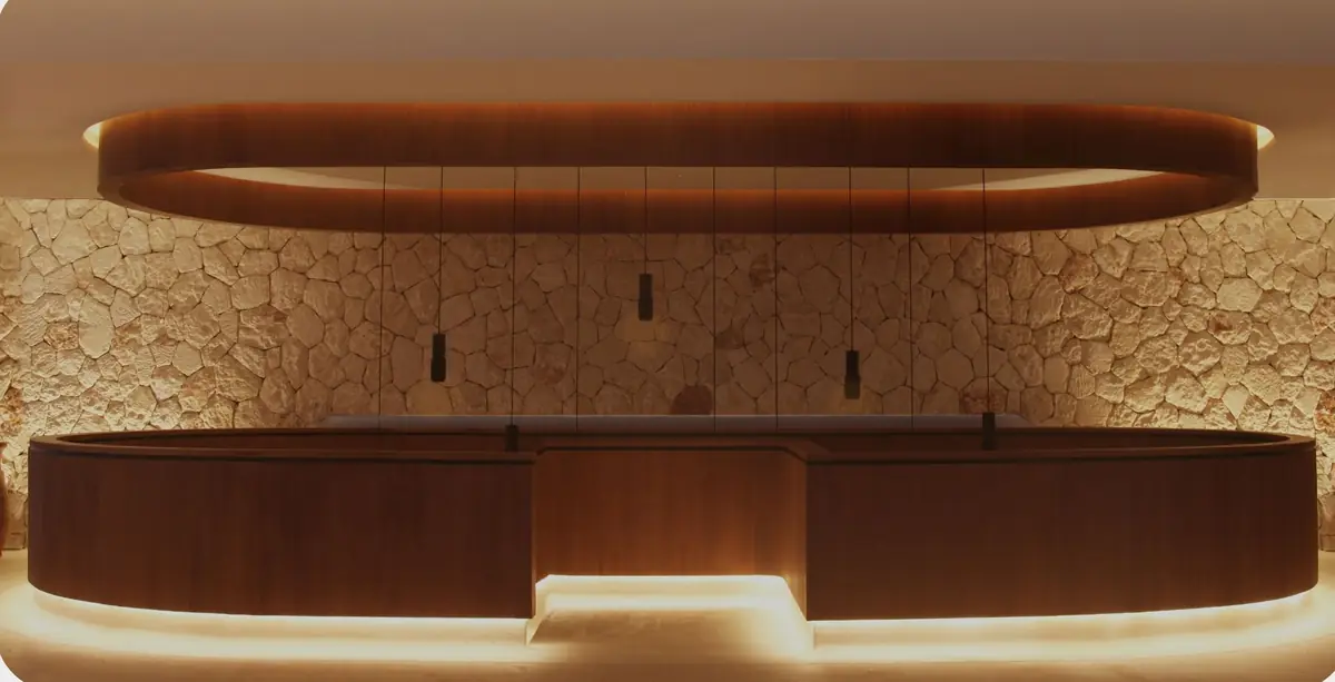 Mostrador de recepción curvo de madera iluminado ante un muro de piedra mallorquina