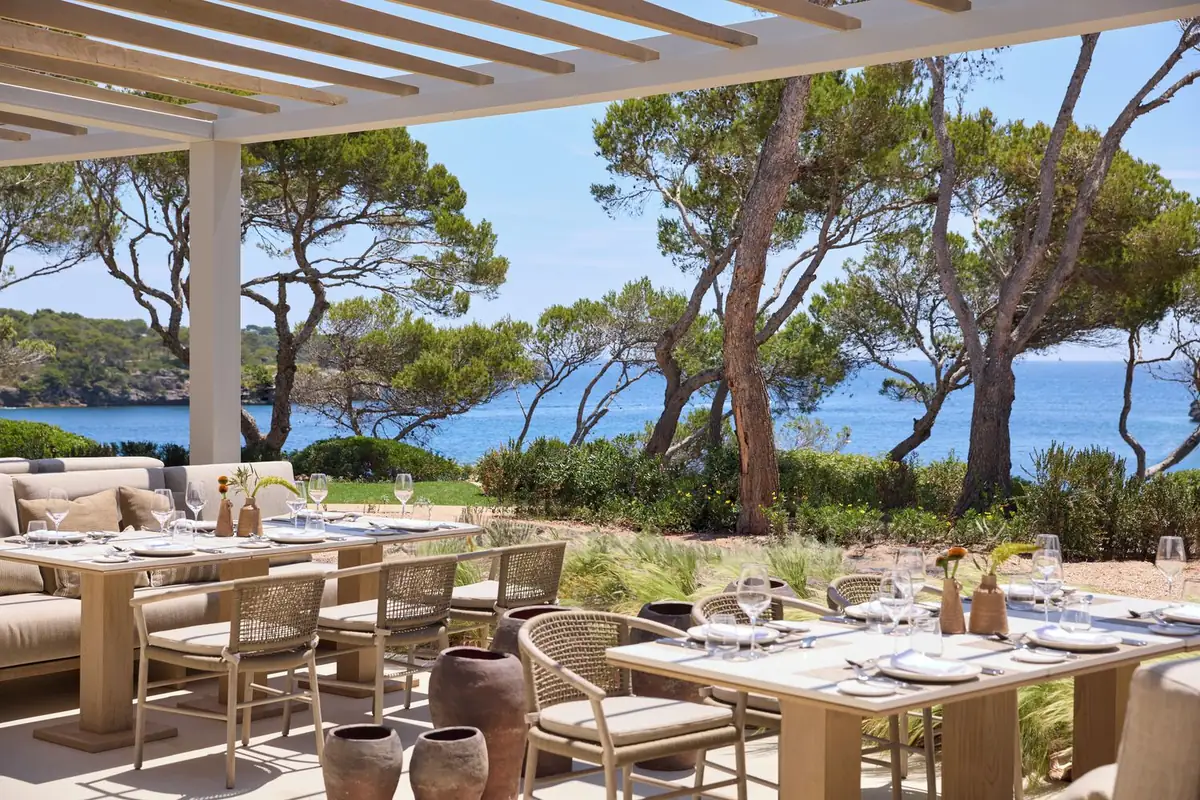 Terraza de restaurante bajo pérgola con mesas de madera, pinos y el mar al fondo