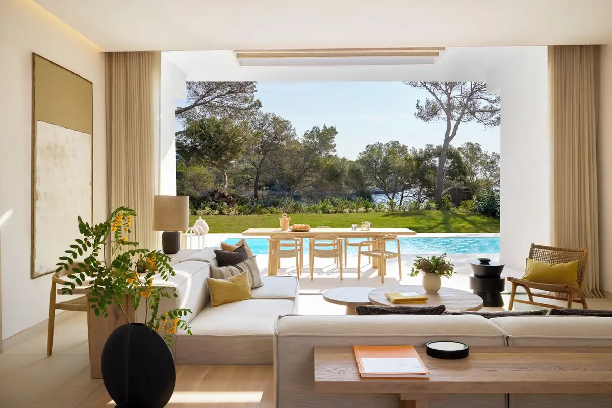 Salón de un bungalow con sofá, butacas de madera y ventanal enmarcando la piscina y los pinos