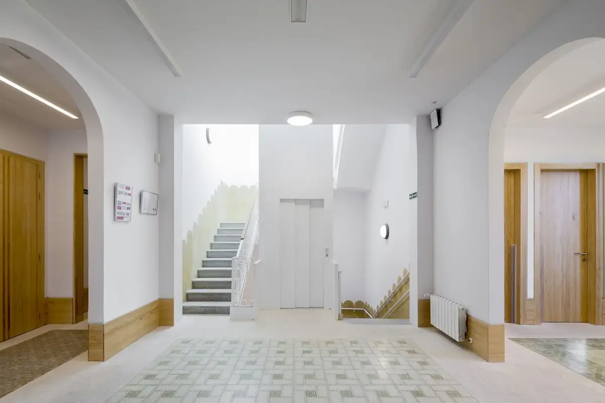 Vestíbulo interior con arcos, escalera, ascensor, zócalo de madera y pavimento de baldosa
