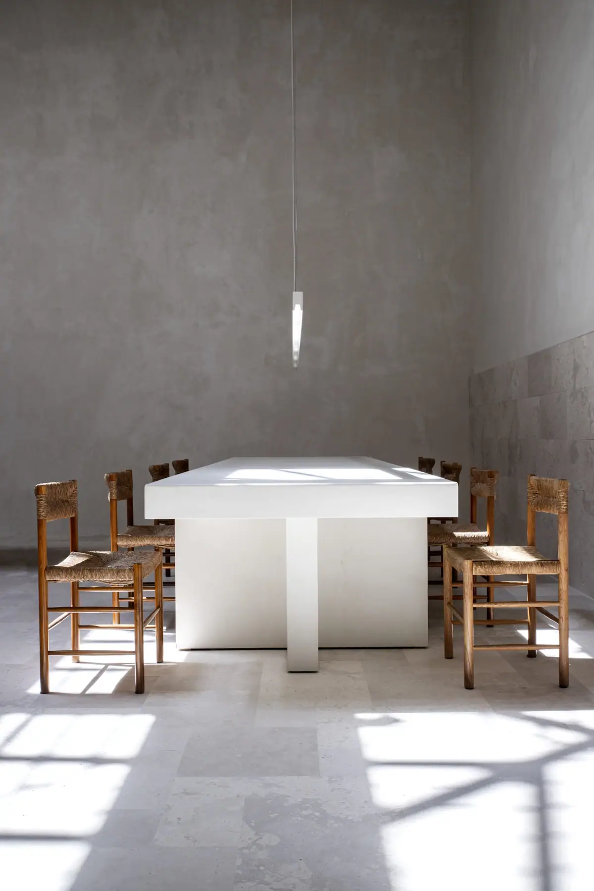White meeting table with wooden chairs with rush seats beneath a pendant lamp, with sunlight falling on the stone floor