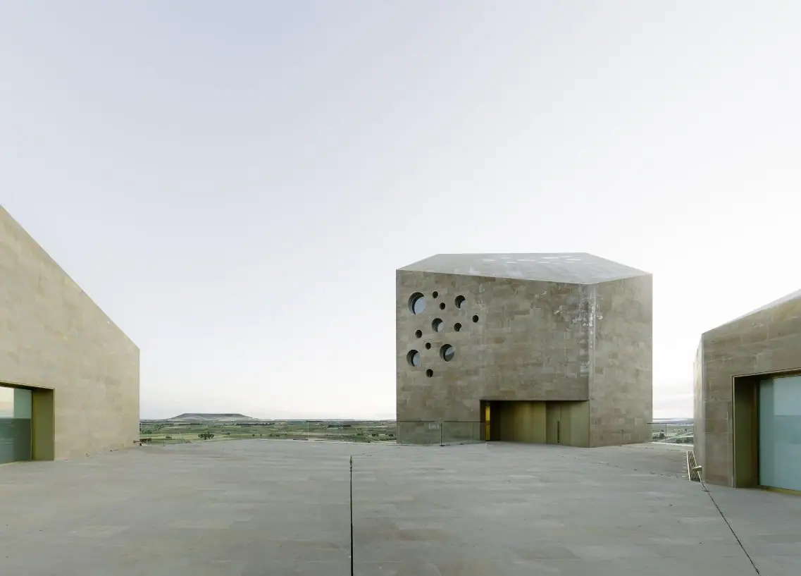 Stone viewing square between ashlar volumes, with the tower of circular openings and the plateau in the background