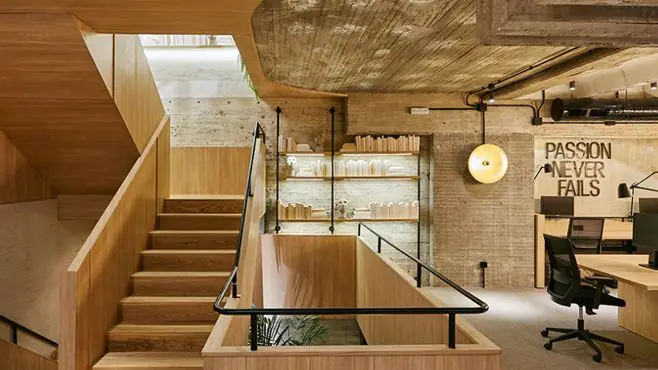 Escalera de madera con barandilla negra junto a una estantería sobre muro de hormigón visto y puestos de trabajo