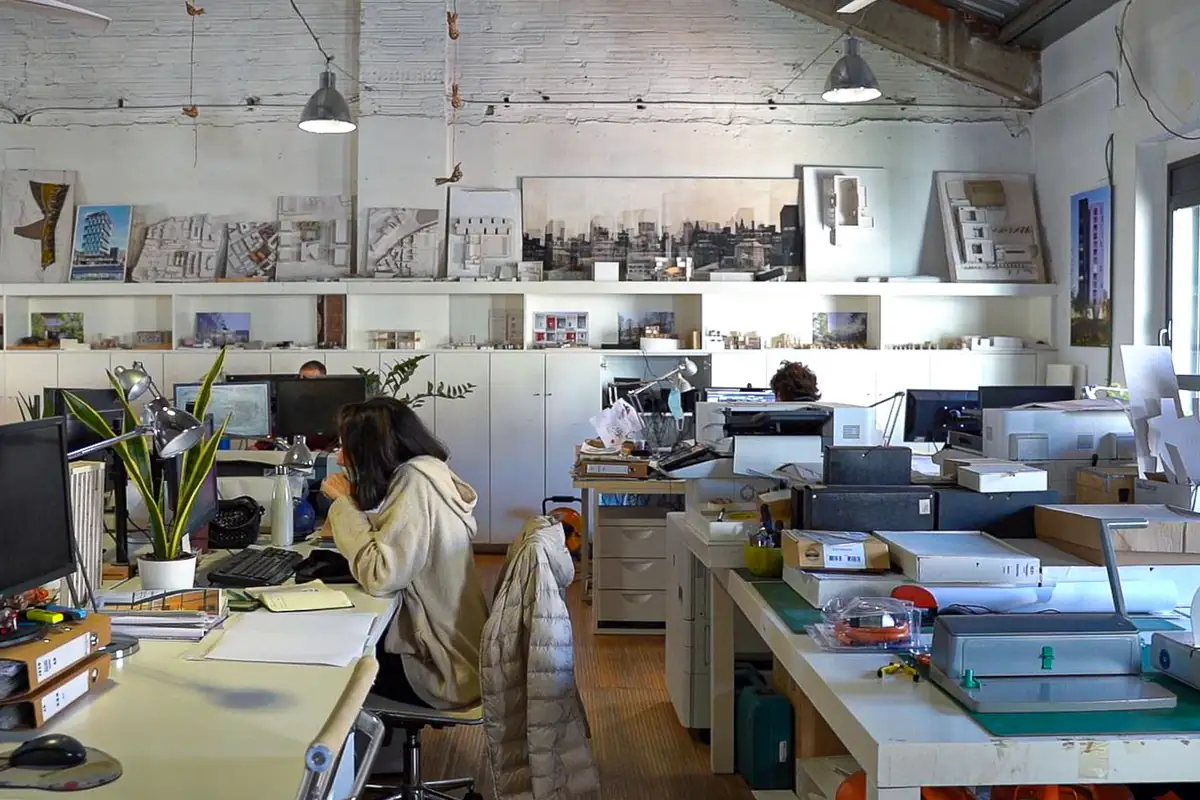 Mesas de trabajo con ordenadores y maquetas en el estudio, bajo una pared de ladrillo pintada de blanco