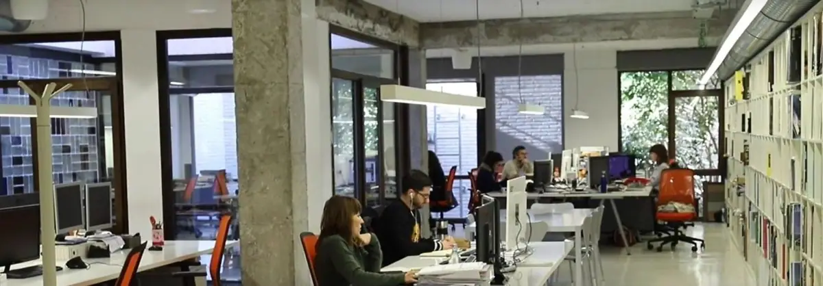 Workstations on an open-plan floor with concrete columns, pendant lamps and a long bookcase