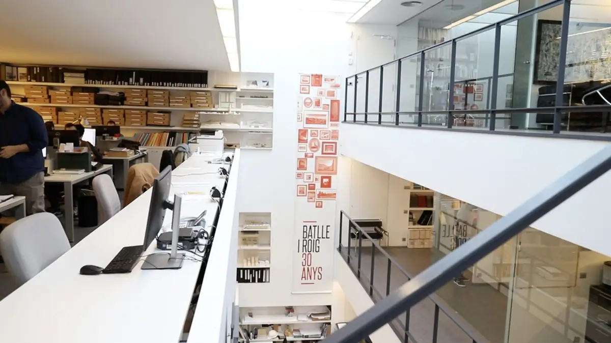 The studio's work floor with long desks next to a double-height space and a glass balustrade