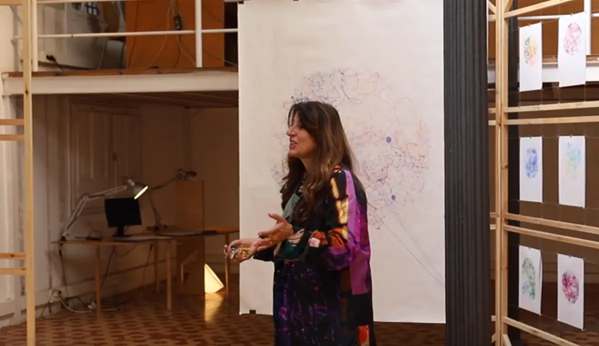 Benedetta Tagliabue speaks standing in front of a large hanging drawing and a wooden slatted structure holding drawings