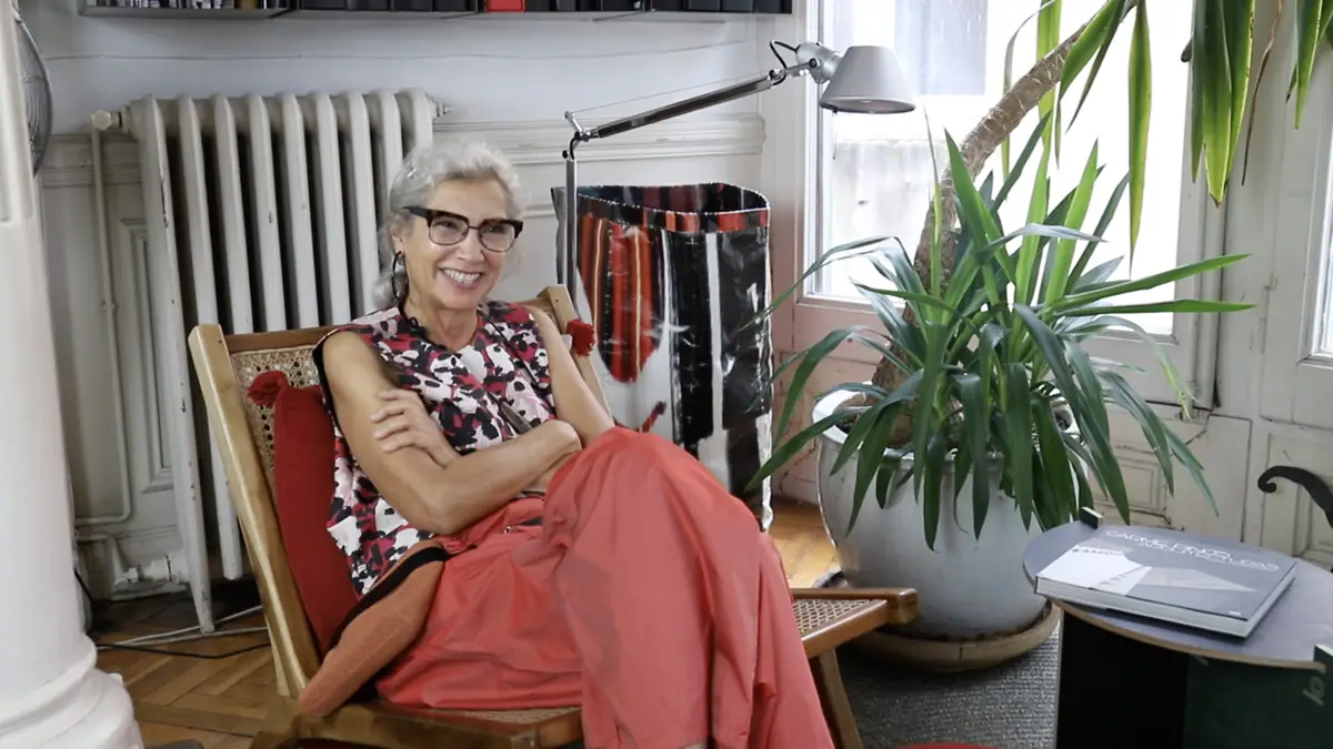 Carme Pinós smiles, seated in an armchair next to a plant and a radiator in her studio