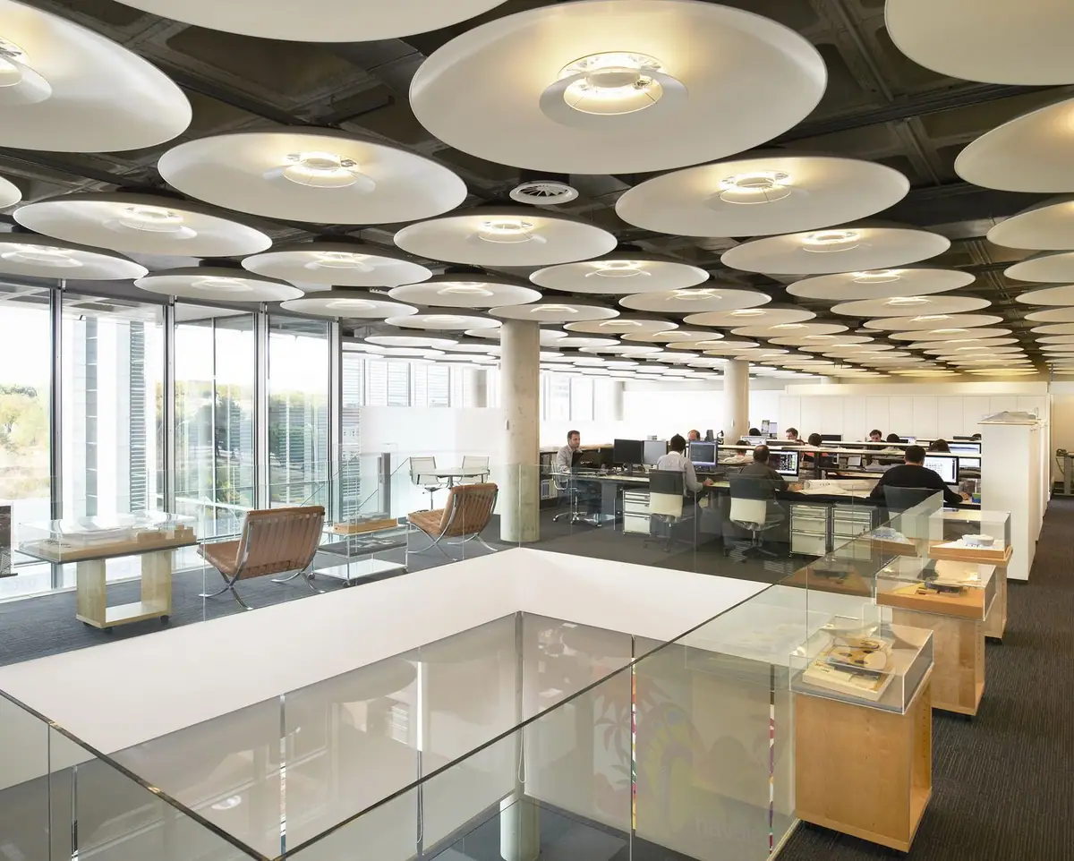 Planta de oficinas con lámparas circulares en el techo, sillones de cuero y vitrinas con maquetas junto a un hueco de doble altura