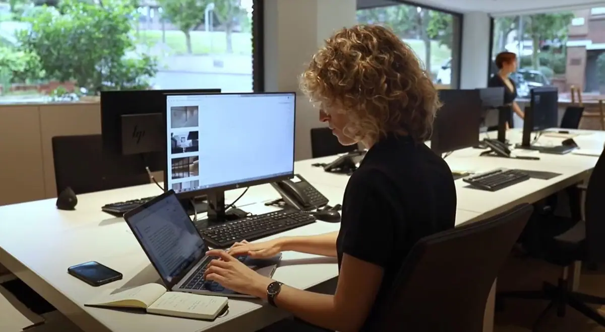 A team member works on a laptop at a long desk in the studio, next to large windows
