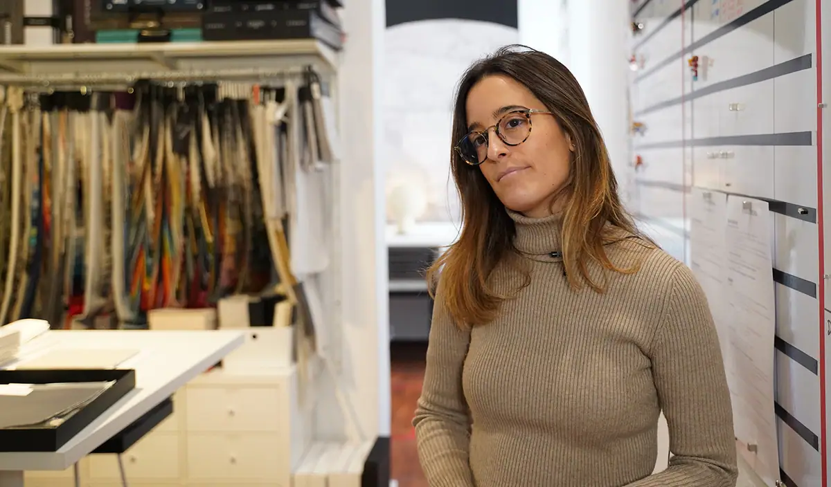 Anna Fajertag, wearing glasses and a roll-neck jumper, beside a fabric sample display in the studio