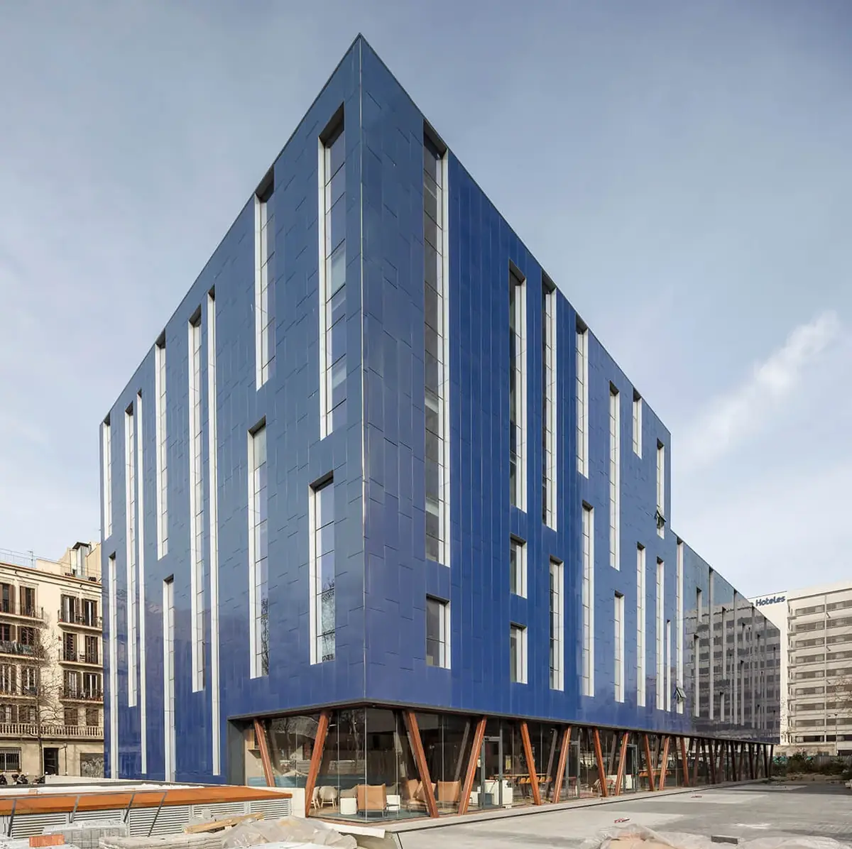 Façade of the Hotel Paxton in Barcelona, clad in vertical pieces in blue tones, with a glazed ground floor on V-shaped columns