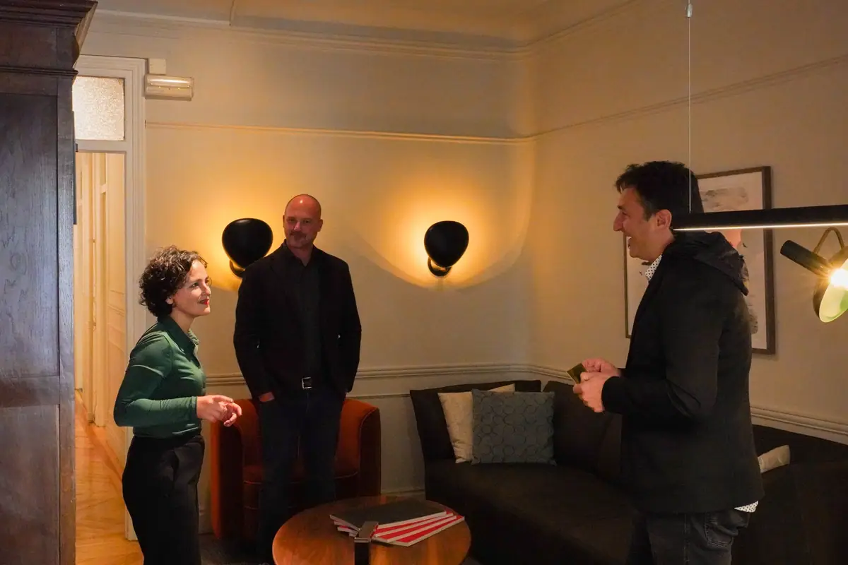 Three people talking, standing in a room with mouldings, black wall lights, a dark sofa and an orange armchair