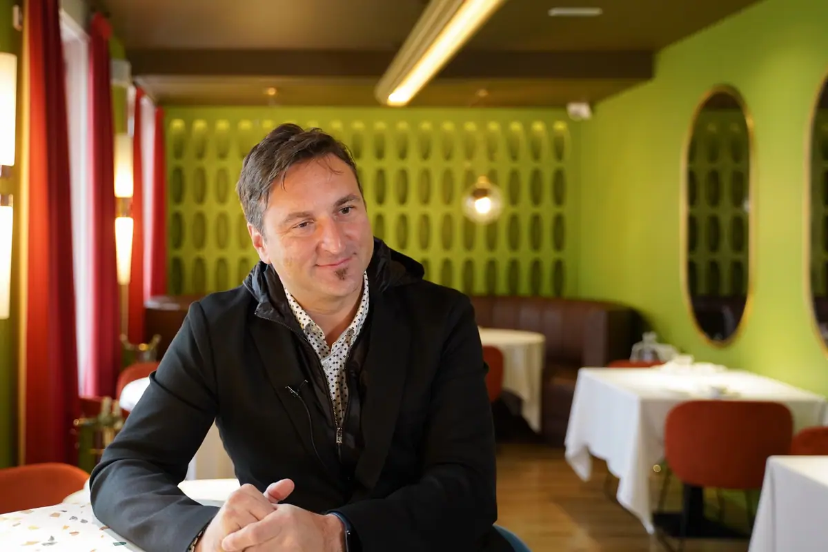 Andrea Spada smiles, seated at a table in a restaurant with green walls, oval mirrors and red chairs
