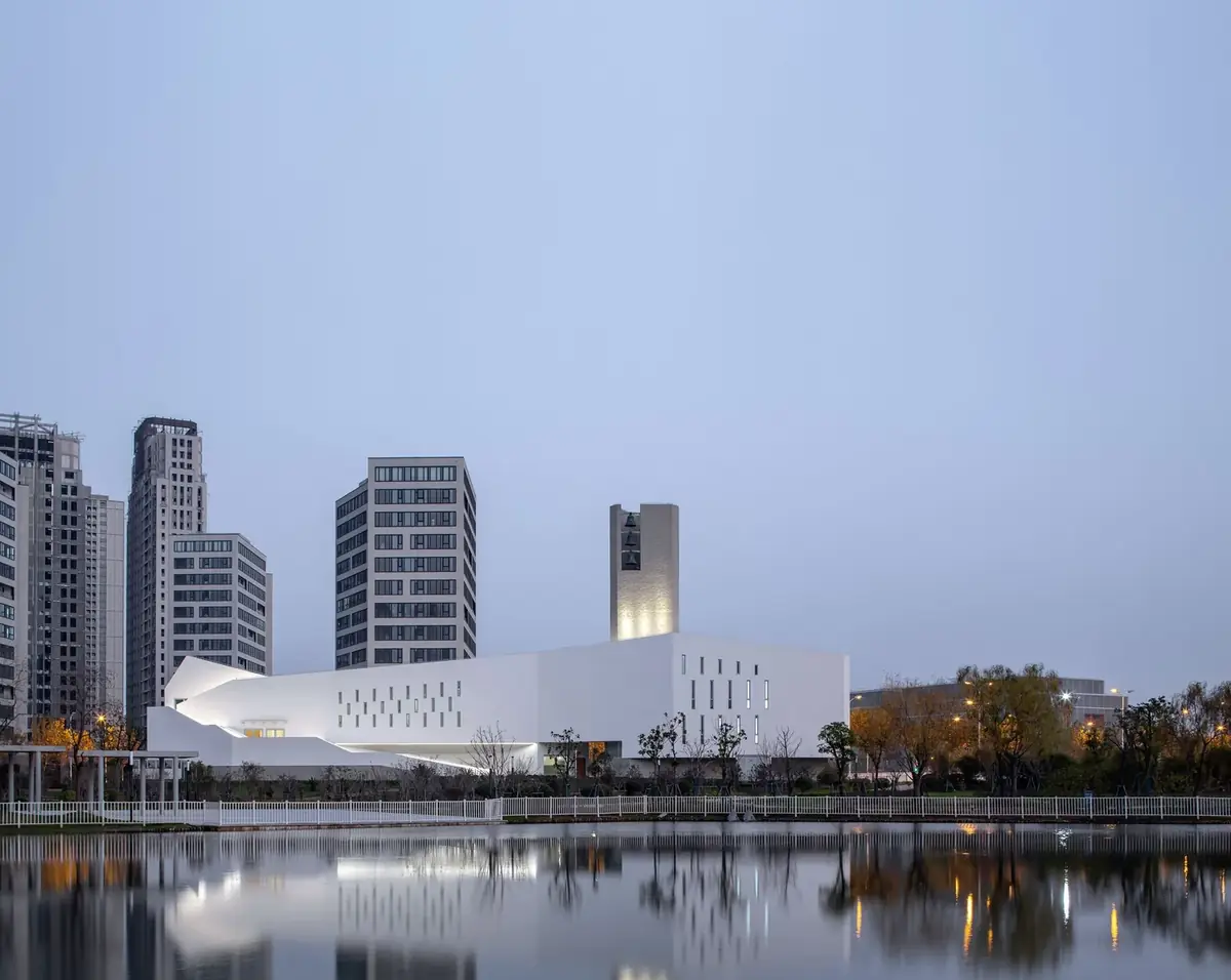 White elongated church with a bell tower reflected in water at dusk, with residential towers behind